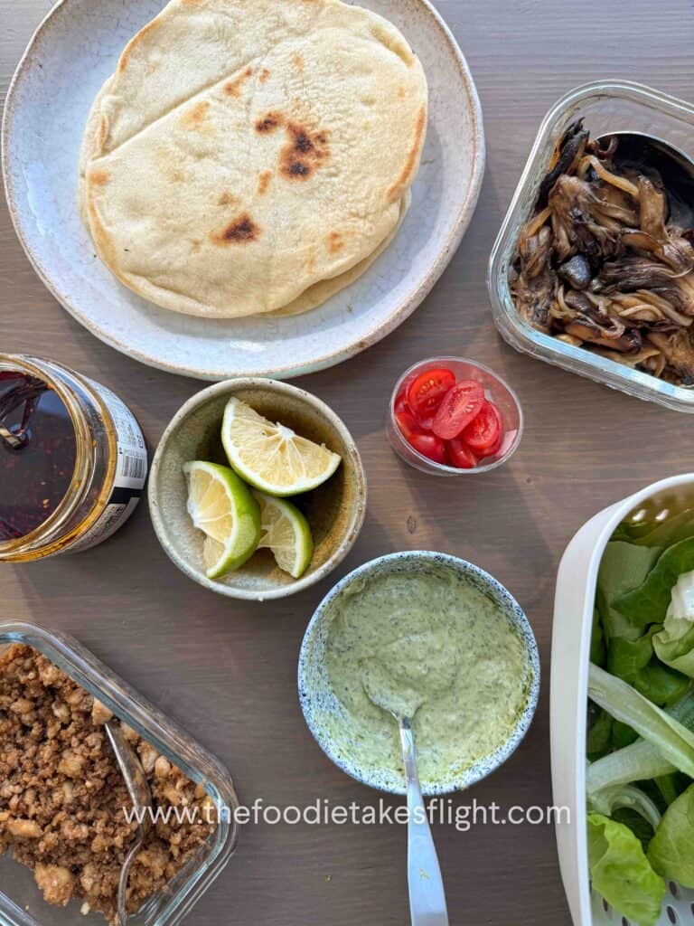 Flat lay of ingredients for tofu and mushroom wraps, including crumbled tofu, sliced mushrooms, pesto mayo, lime, and fresh vegetables