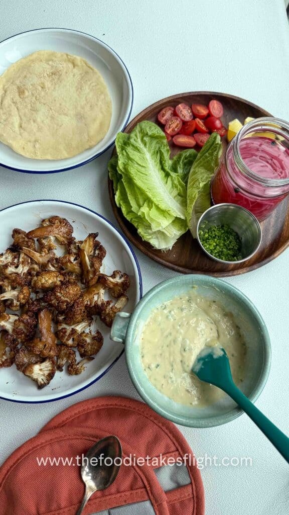 Bowls of roasted cauliflower, garlic mayo, pickled onions, and fresh veggies arranged for assembling wraps