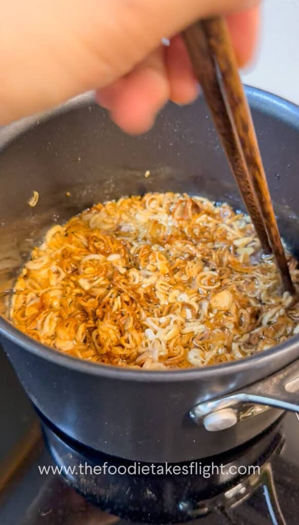 Thinly sliced shallots gently frying in neutral oil in a pot, bubbling as they crisp