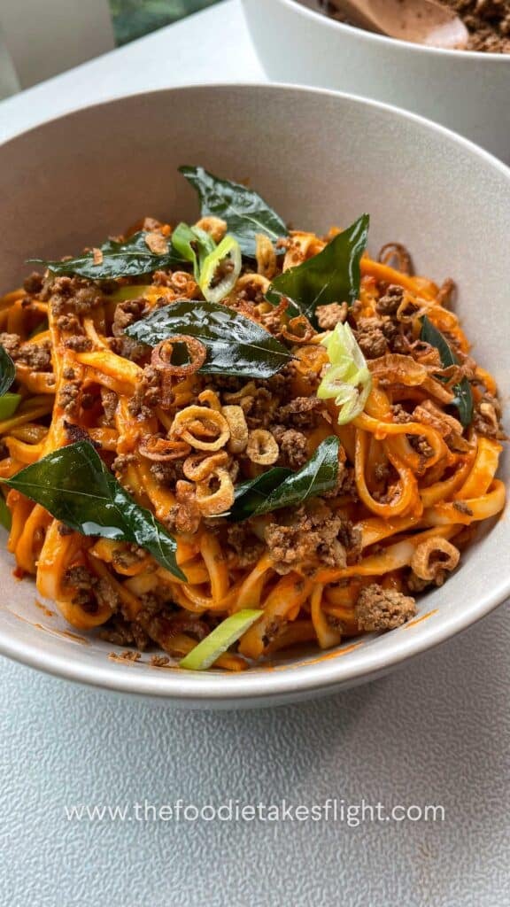Close up of creamy tomato coconut noodles topped with fried curry leaves, crumbled tofu, and crispy shallots in a white bowl