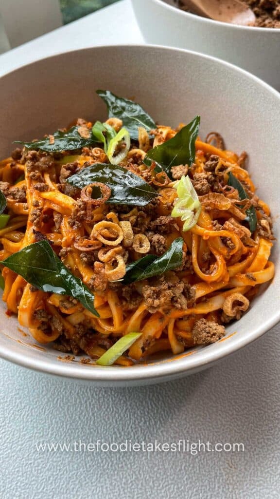 Close up of creamy tomato coconut noodles topped with fried curry leaves, crumbled tofu, and crispy shallots in a white bowl