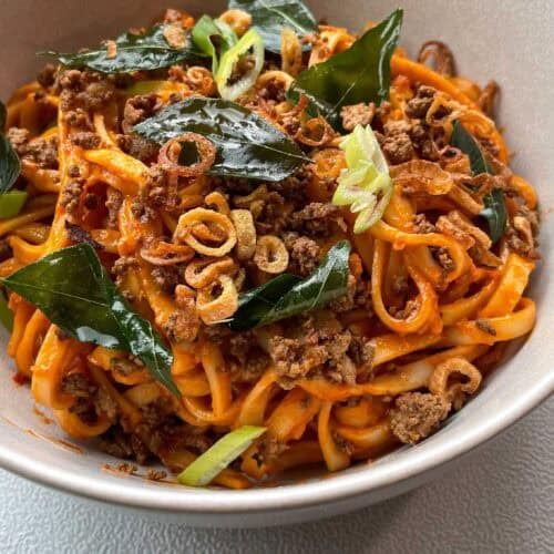 Close up of creamy tomato coconut noodles topped with fried curry leaves, crumbled tofu, and crispy shallots in a white bowl