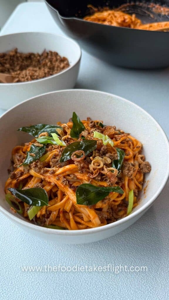 Close up of creamy tomato coconut noodles topped with fried curry leaves, crumbled tofu, and crispy shallots in a white bowl