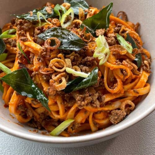 Close up of creamy tomato coconut noodles topped with fried curry leaves, crumbled tofu, and crispy shallots in a white bowl