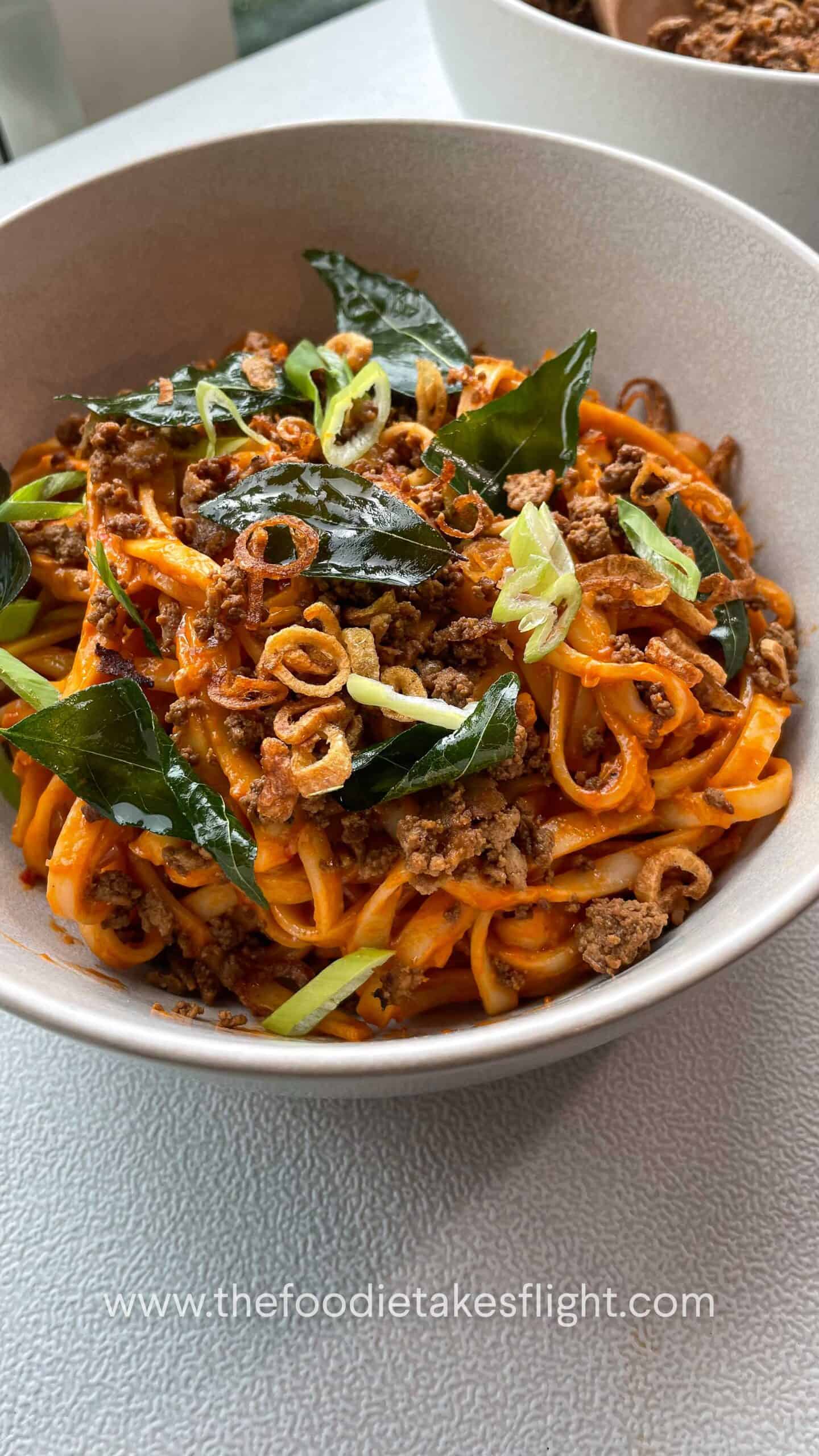Close up of creamy tomato coconut noodles topped with fried curry leaves, crumbled tofu, and crispy shallots in a white bowl