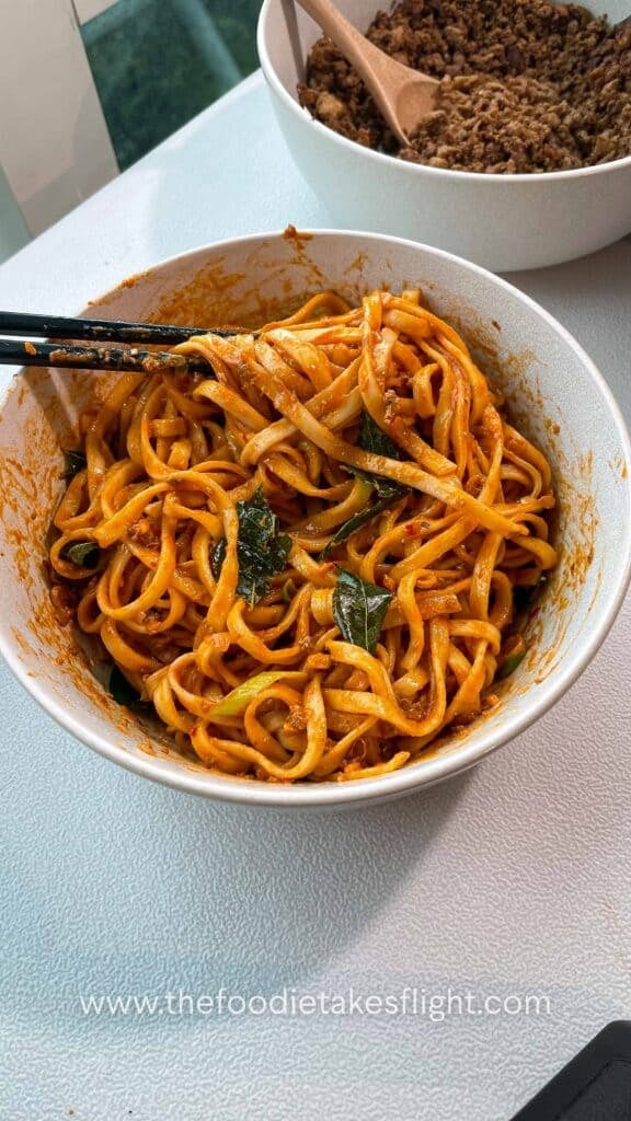 Close up of creamy tomato coconut noodles topped with fried curry leaves, crumbled tofu, and crispy shallots in a white bowl