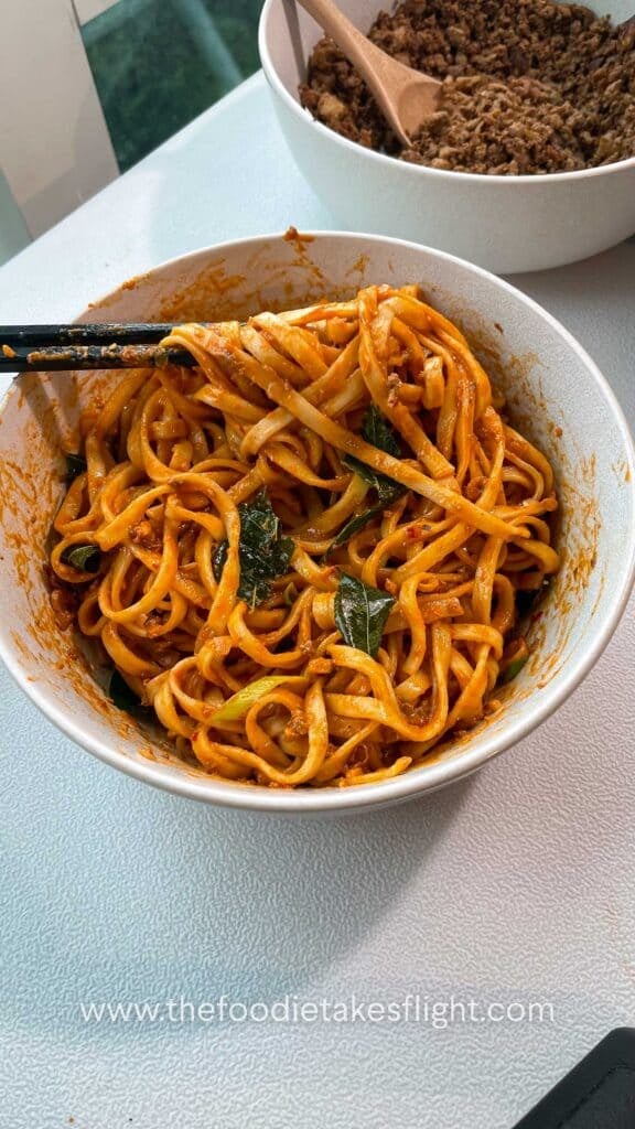 Close up of creamy tomato coconut noodles topped with fried curry leaves, crumbled tofu, and crispy shallots in a white bowl