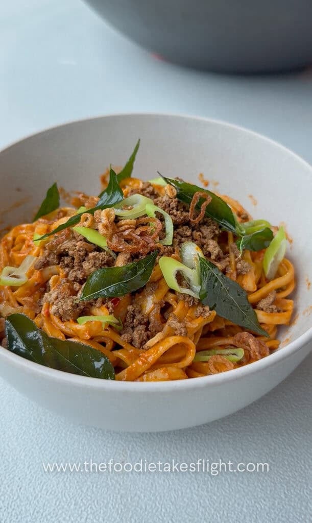 Close up of creamy tomato coconut noodles topped with fried curry leaves, crumbled tofu, and crispy shallots in a white bowl