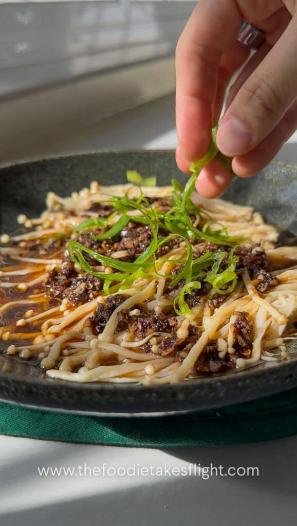 Steamed enoki mushrooms being garnished with sliced green onions and sesame oil