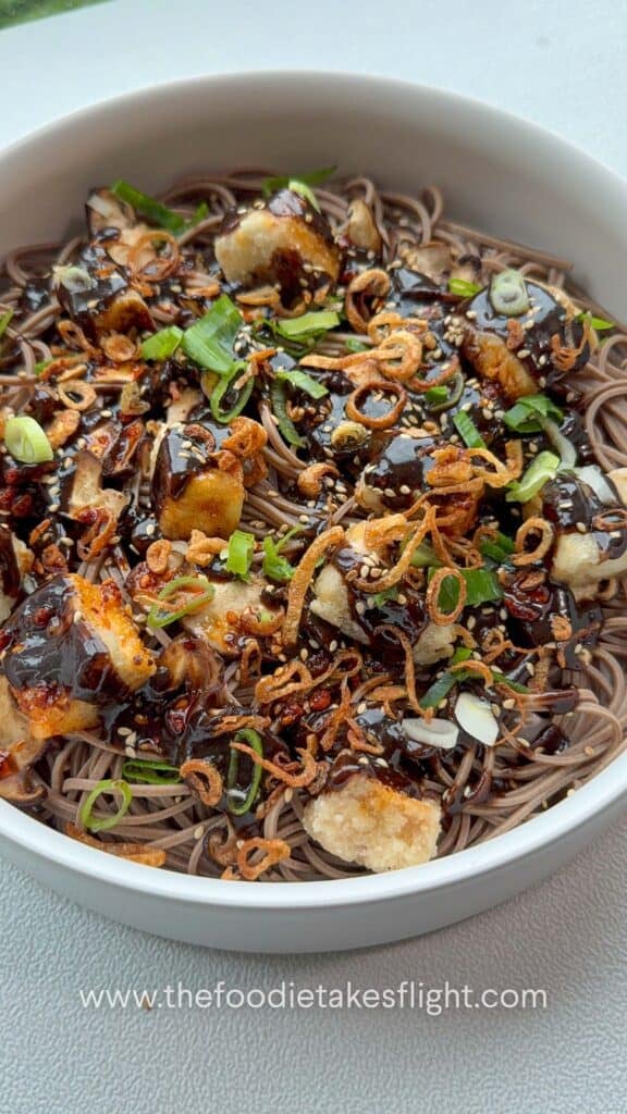 Bowl of soba noodles with black garlic miso dressing, topped with crispy tofu, mushrooms, and fried shallots.