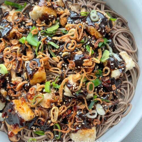 Bowl of soba noodles with black garlic miso dressing, topped with crispy tofu, mushrooms, and fried shallots.
