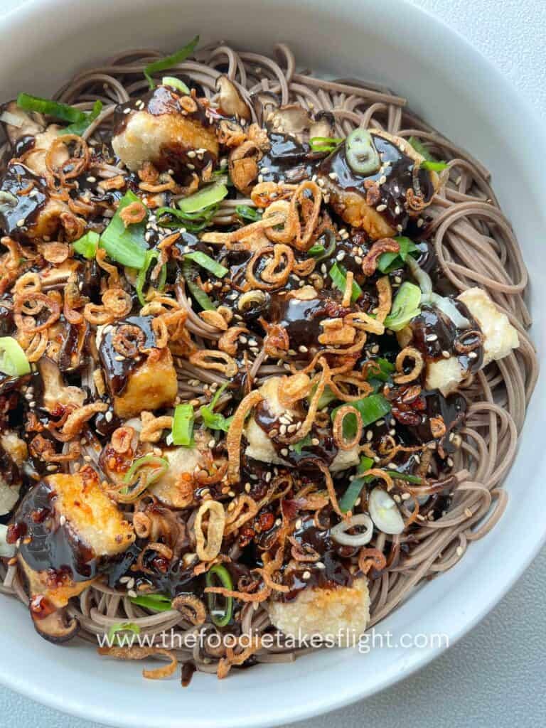 Bowl of soba noodles with black garlic miso dressing, topped with crispy tofu, mushrooms, and fried shallots.