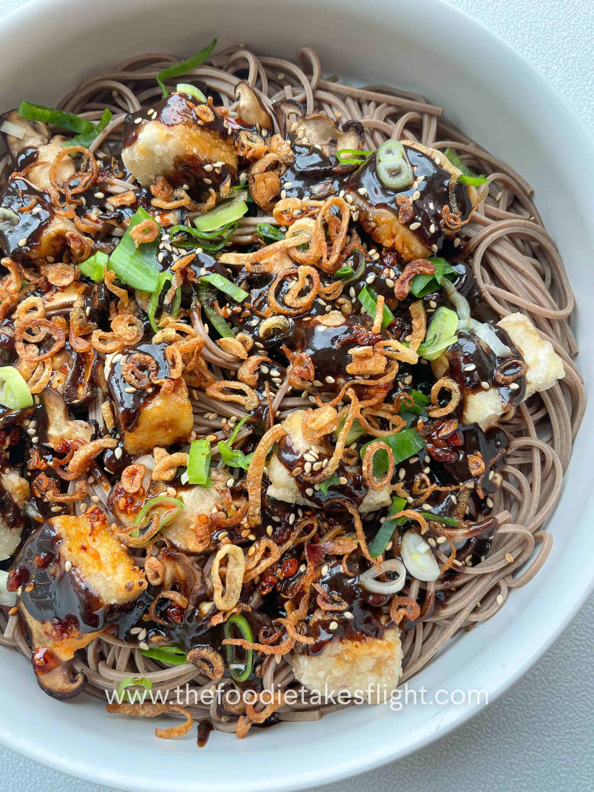 Bowl of soba noodles with black garlic miso dressing, topped with crispy tofu, mushrooms, and fried shallots.