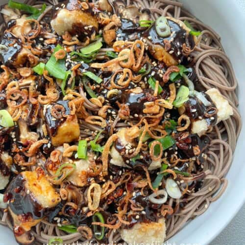 Bowl of soba noodles with black garlic miso dressing, topped with crispy tofu, mushrooms, and fried shallots.