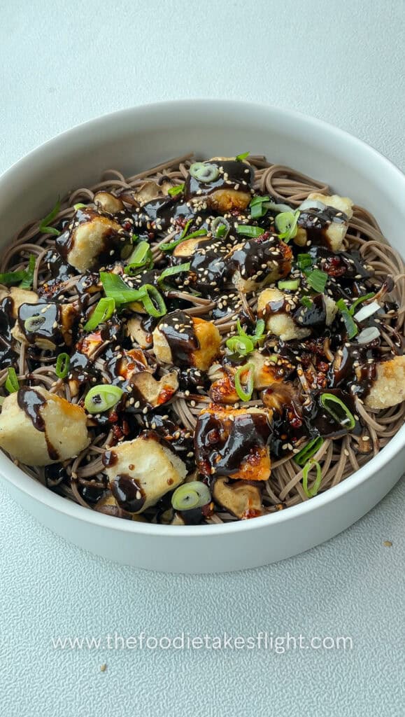 Bowl of soba noodles tossed in black garlic miso dressing, topped with crispy tofu, mushrooms, and fried shallots