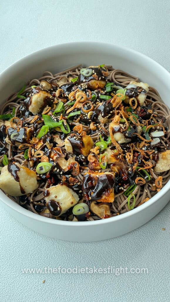 Bowl of soba noodles tossed in black garlic miso dressing, topped with crispy tofu, mushrooms, and fried shallots