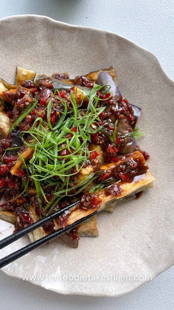 Bowl of Chinese fried eggplant with spicy garlic sauce, with small red bell pepper pieces and thinly sliced green onions
