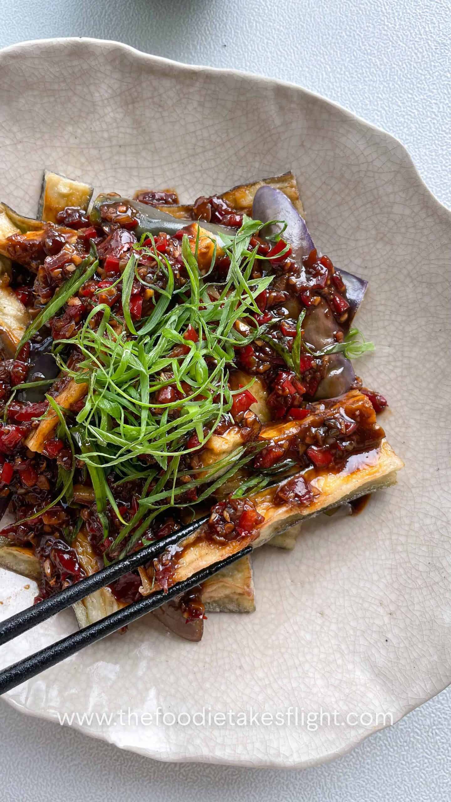 Bowl of Chinese fried eggplant with spicy garlic sauce, with small red bell pepper pieces and thinly sliced green onions