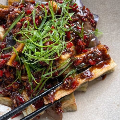 Bowl of Chinese fried eggplant with spicy garlic sauce, with small red bell pepper pieces and thinly sliced green onions