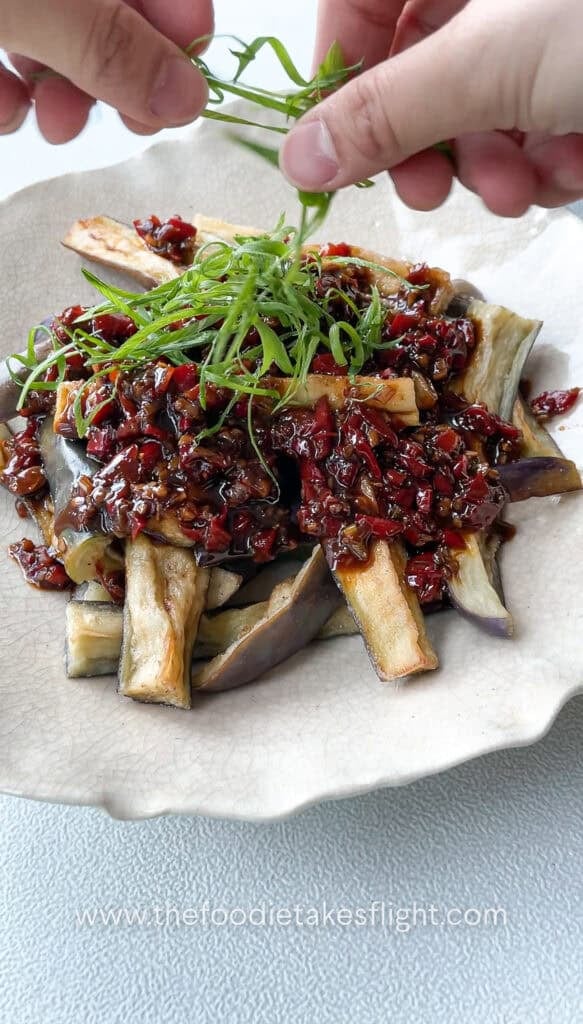 Bowl of Chinese fried eggplant with spicy garlic sauce, with small red bell pepper pieces and thinly sliced green onions