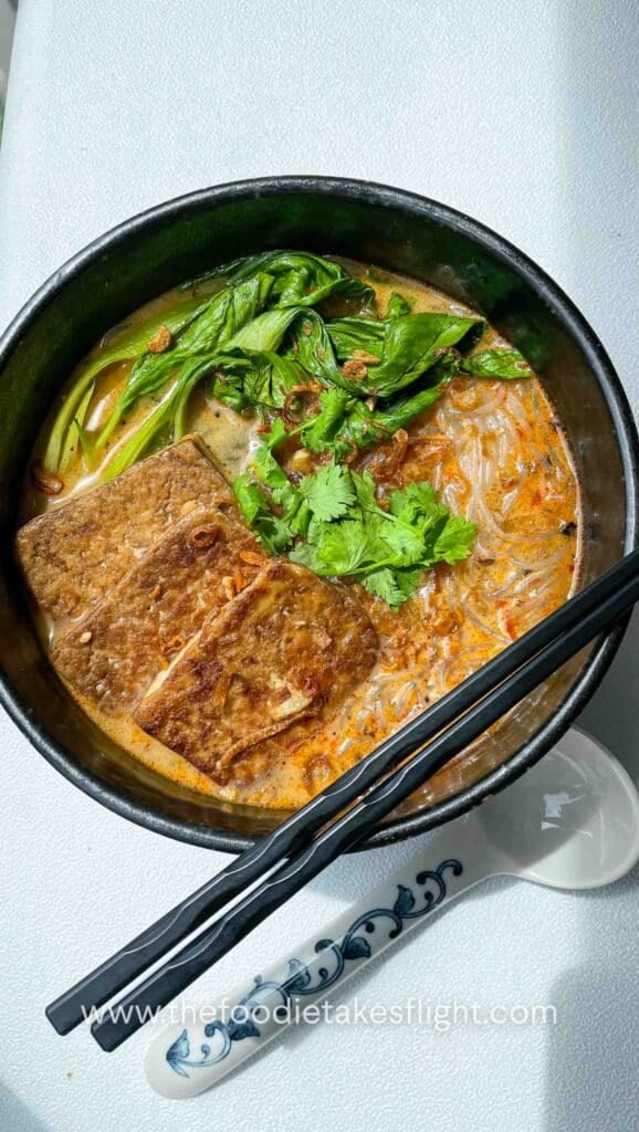 Bowl of spicy lemongrass coconut noodle soup topped with bok choy, tofu cubes, cilantro, and fried shallots