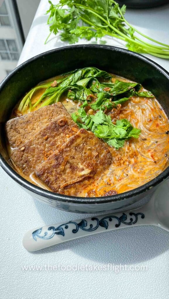 Bowl of spicy lemongrass coconut noodle soup topped with bok choy, tofu cubes, cilantro, and fried shallots