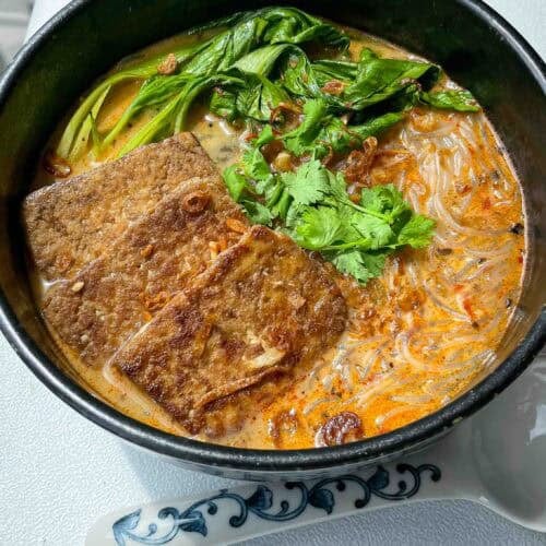 Bowl of spicy lemongrass coconut noodle soup topped with bok choy, tofu cubes, cilantro, and fried shallots