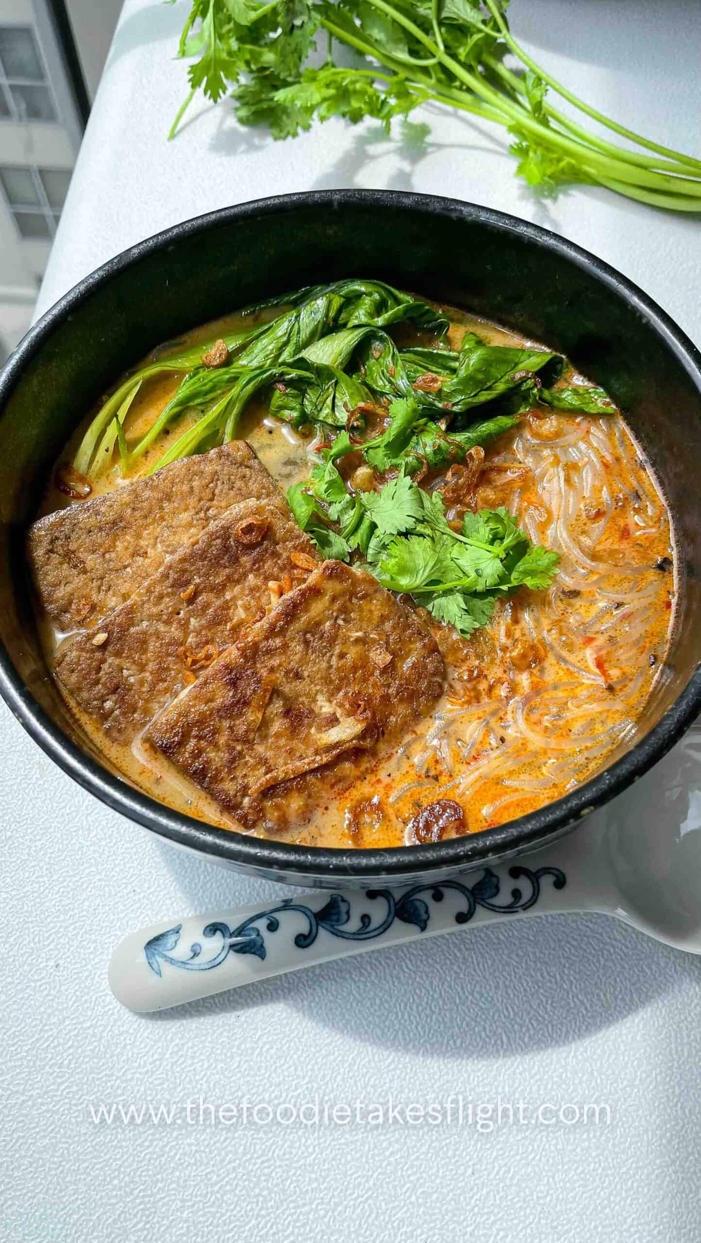 Bowl of spicy lemongrass coconut noodle soup topped with bok choy, tofu cubes, cilantro, and fried shallots