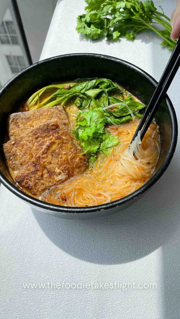Bowl of spicy lemongrass coconut noodle soup topped with bok choy, tofu cubes, cilantro, and fried shallots