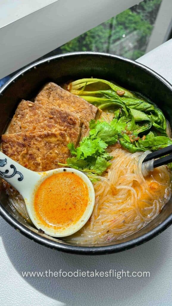 Bowl of spicy lemongrass coconut noodle soup topped with bok choy, tofu cubes, cilantro, and fried shallots