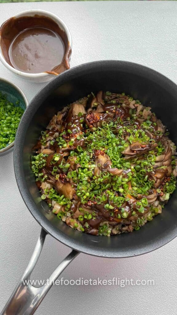 Overhead shot of a rice pot filled with glazed maitake mushrooms, drizzles of black garlic miso sauce and chives