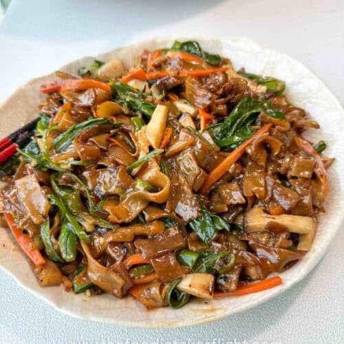 Bowl of stir-fried hor fun rice noodles topped with vegetables, gravy, fried shallots, and chili oil.