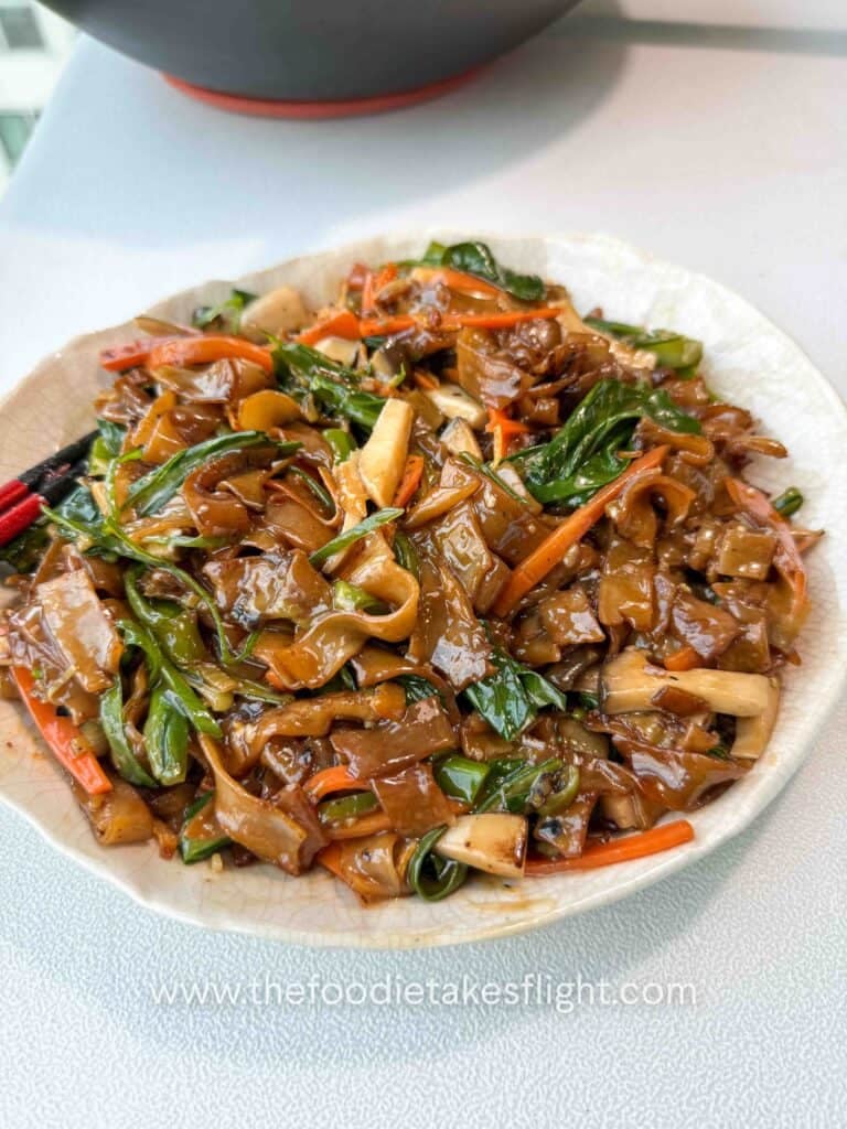 Bowl of stir-fried hor fun rice noodles topped with vegetables, gravy, fried shallots, and chili oil.