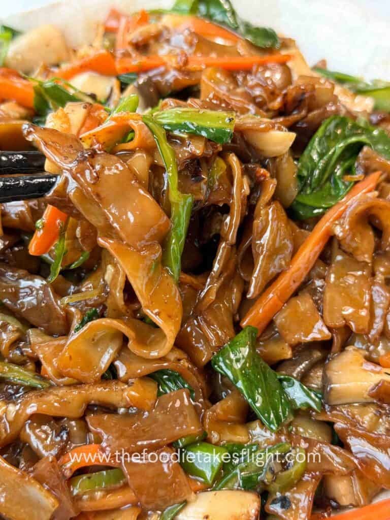 Bowl of stir-fried hor fun rice noodles topped with vegetables, gravy, fried shallots, and chili oil.