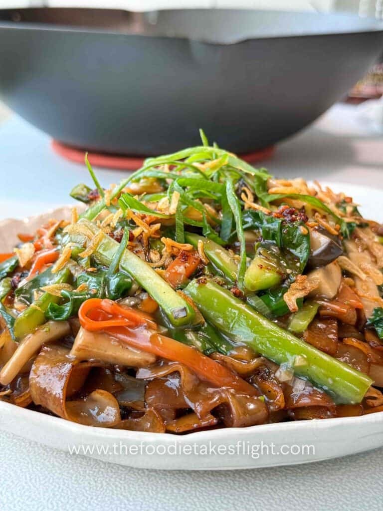 Bowl of stir-fried hor fun rice noodles topped with vegetables, gravy, fried shallots, and chili oil.
