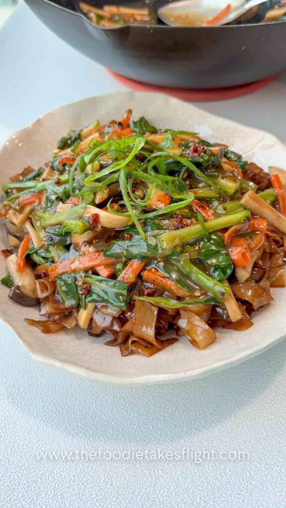 Bowl of stir-fried hor fun rice noodles topped with vegetables, gravy, fried shallots, and chili oil.