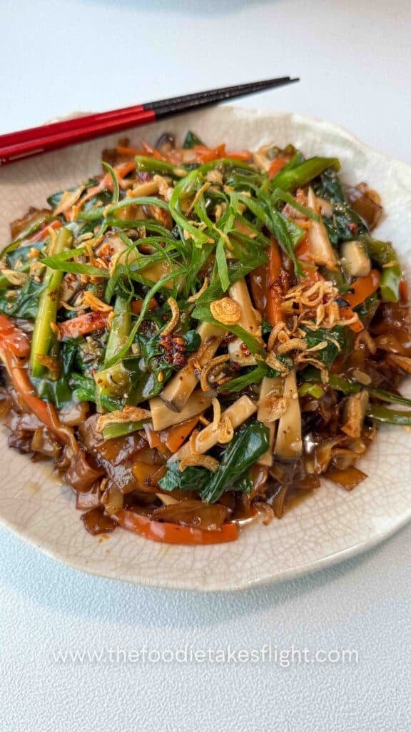 Bowl of stir-fried hor fun rice noodles topped with vegetables, gravy, fried shallots, and chili oil.