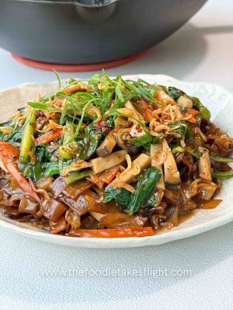 Bowl of stir-fried hor fun rice noodles topped with vegetables, gravy, fried shallots, and chili oil.