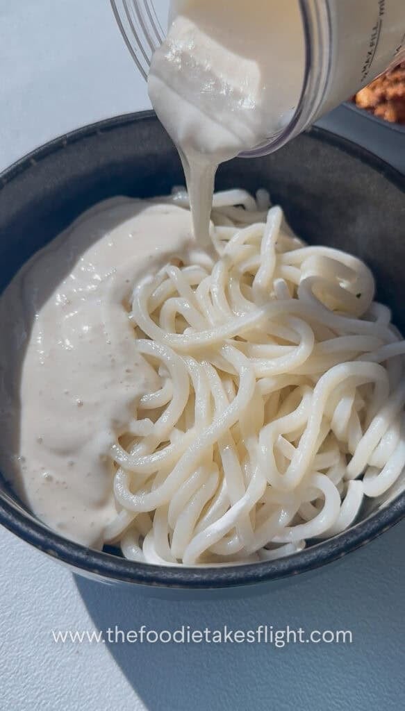 pouring silken tofu sauce over udon noodles in a bowl