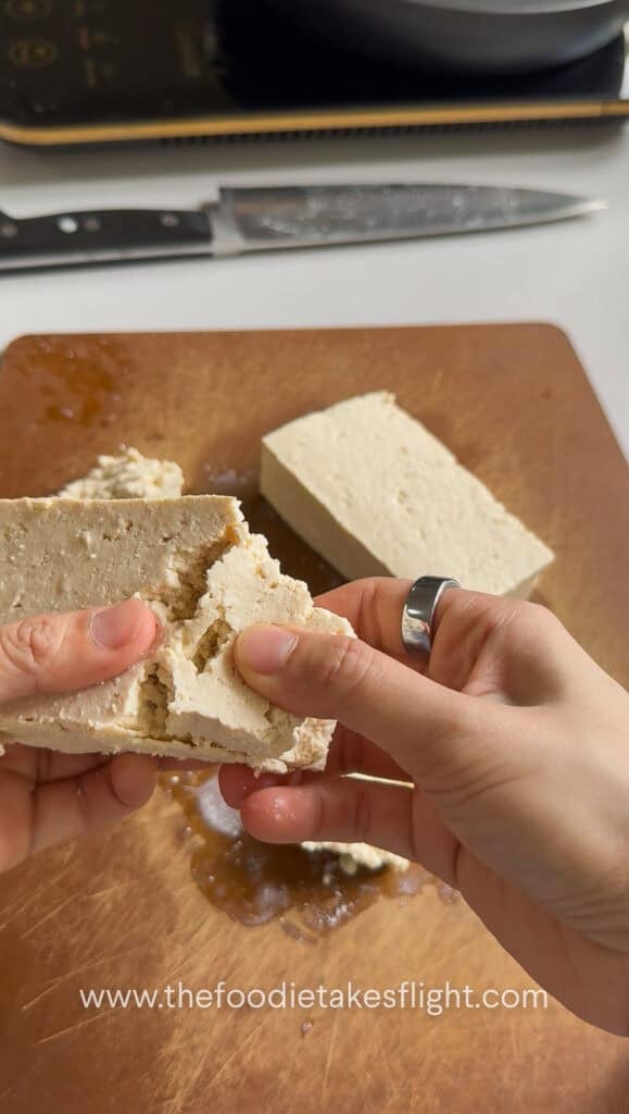 breaking apart a block of extra firm tofu on a wooden cutting board